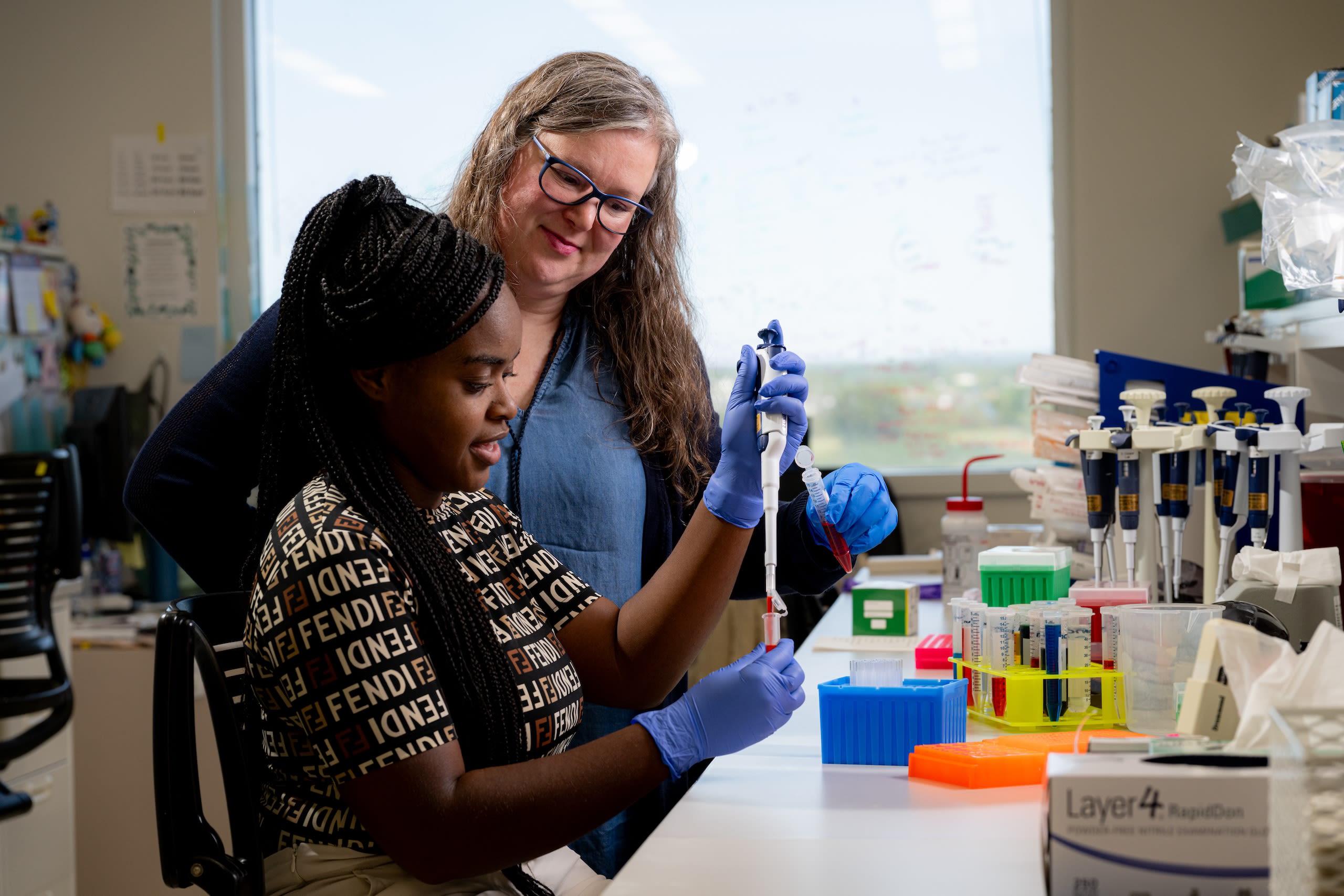 A scientist mentoring a younger scientist who is using a pipette 